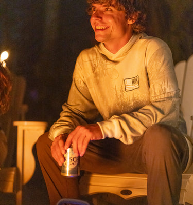 A man sitting outdoors at night by a campfire, wearing a light-colored hoodie with a small chest patch and holding a can. The scene is warmly lit, with dark foliage in the background.