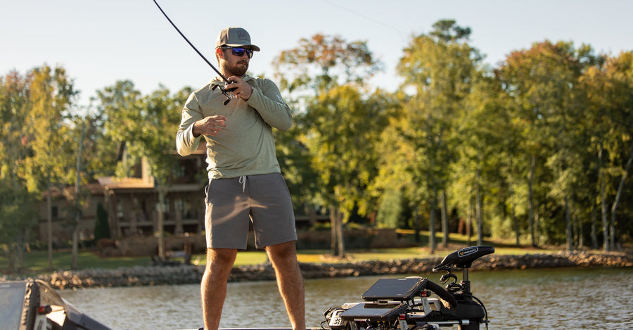 A man stands on a boat on a lake, holding a bent fishing rod. He is wearing a light olive green long-sleeve shirt, gray shorts, and a baseball cap.