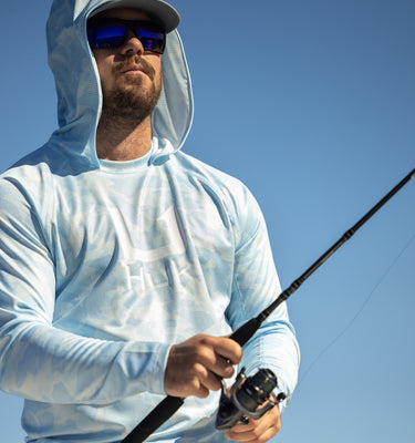 A man wearing a light blue hooded long-sleeve shirt and sunglasses is holding a fishing rod against a clear blue sky.