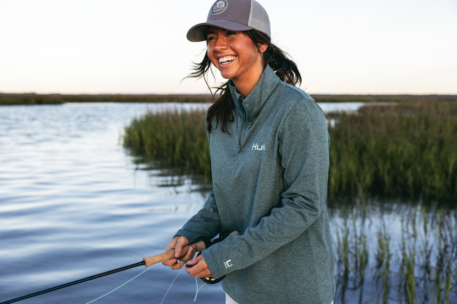 Woman fishing by a lake wearing a blue jacket and cap.