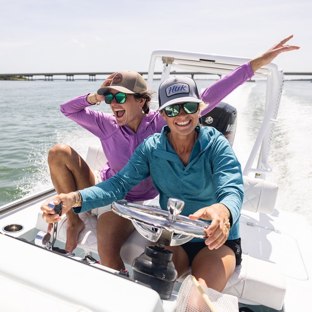 Two women smiling and laughing on a white motorboat moving across the water. The woman in front wears a blue long-sleeve top, black shorts, a gray and blue 'Huk' cap, and sunglasses, while steering the wheel.