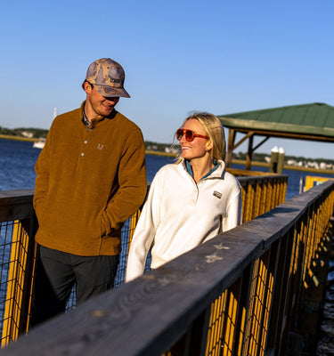 A man and a woman standing on a wooden dock by the water, wearing outdoor apparel. The man wears a brown long-sleeve polo shirt, dark pants, and a camouflage baseball cap.