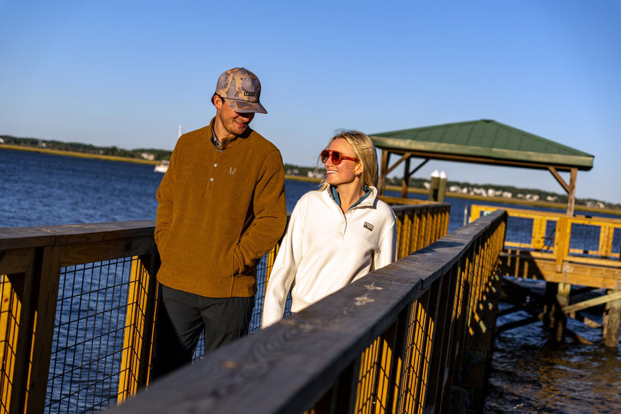 A man and a woman standing on a wooden dock by the water, wearing outdoor apparel. The man wears a brown long-sleeve polo shirt, dark pants, and a camouflage baseball cap.