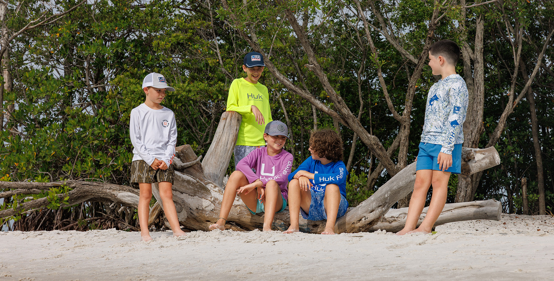 Five children wearing matching branded swim shirts and swim trunks stand and sit on a sandy beach in front of a large piece of driftwood and green trees.
