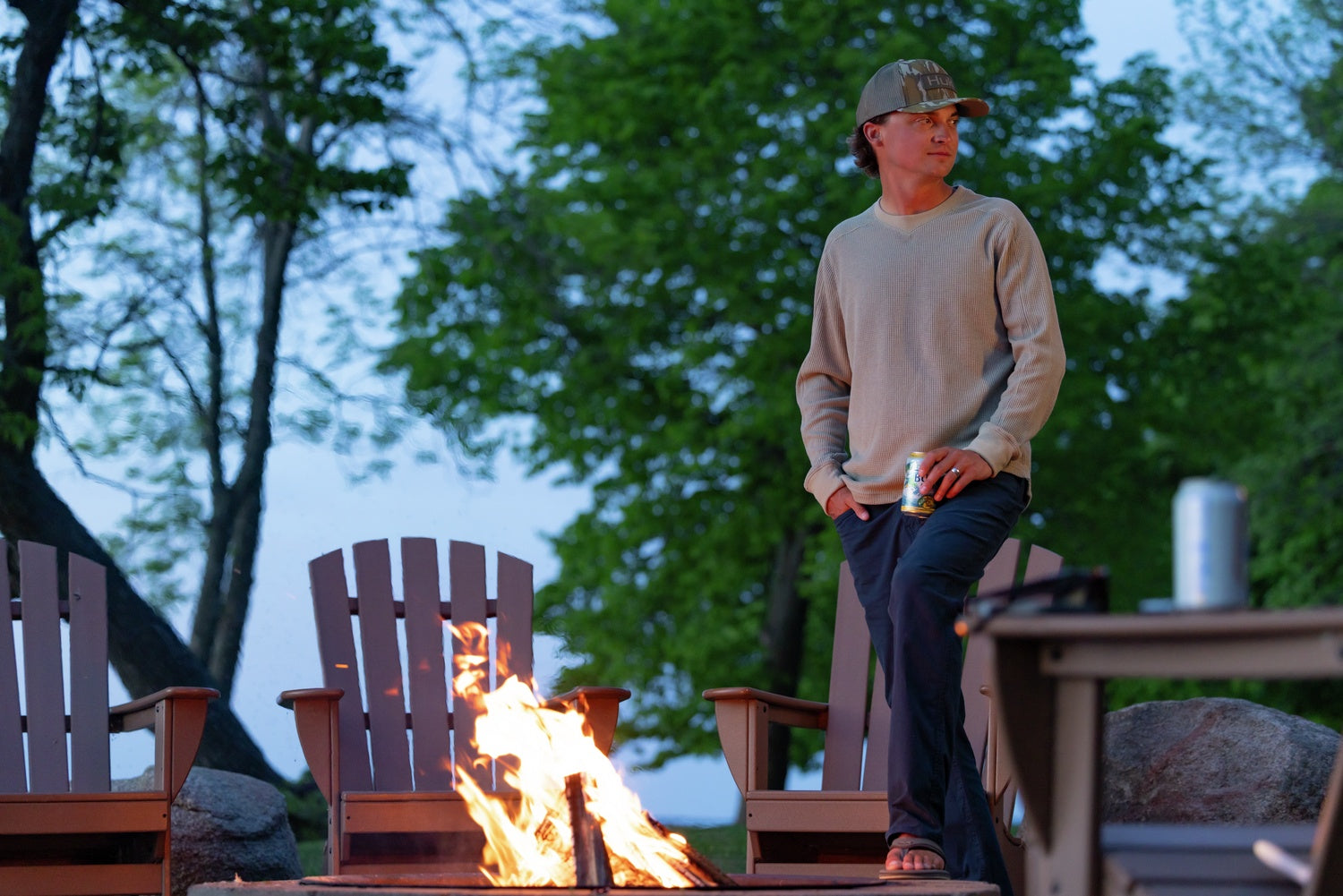 A man wearing a beige long-sleeve shirt, dark pants, and a camouflage cap stands beside a fire pit table in an outdoor patio setting with Adirondack chairs and trees in the background.