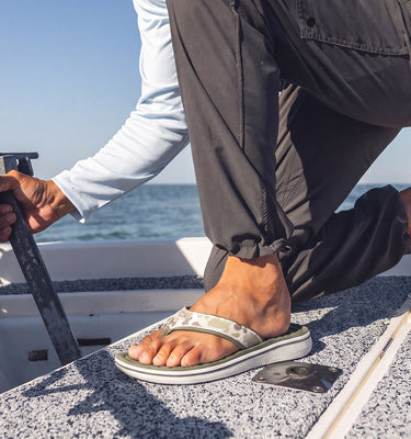 A person crouching on the deck of a boat on sunny days, wearing dark pants, a light long-sleeve shirt, and silver flip-flop sandals with white soles. They are holding a metal tool near the anchor cleat, with the blue ocean and sky in the background.