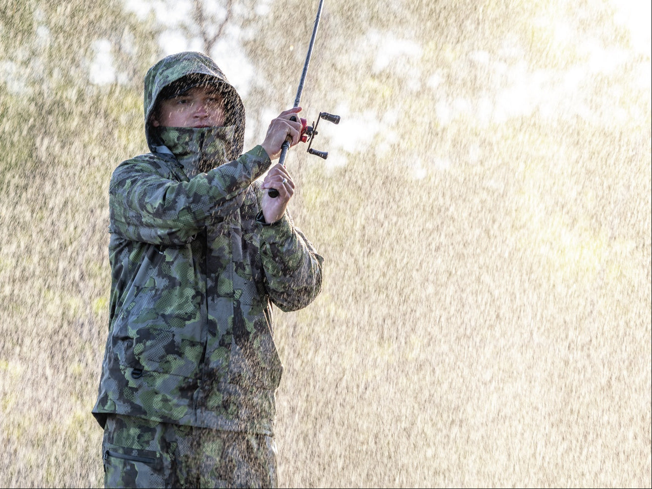 A person wearing a green camouflage hooded rain jacket and matching pants stands outdoors holding a fishing rod, with rain falling and blurred trees in the background.
